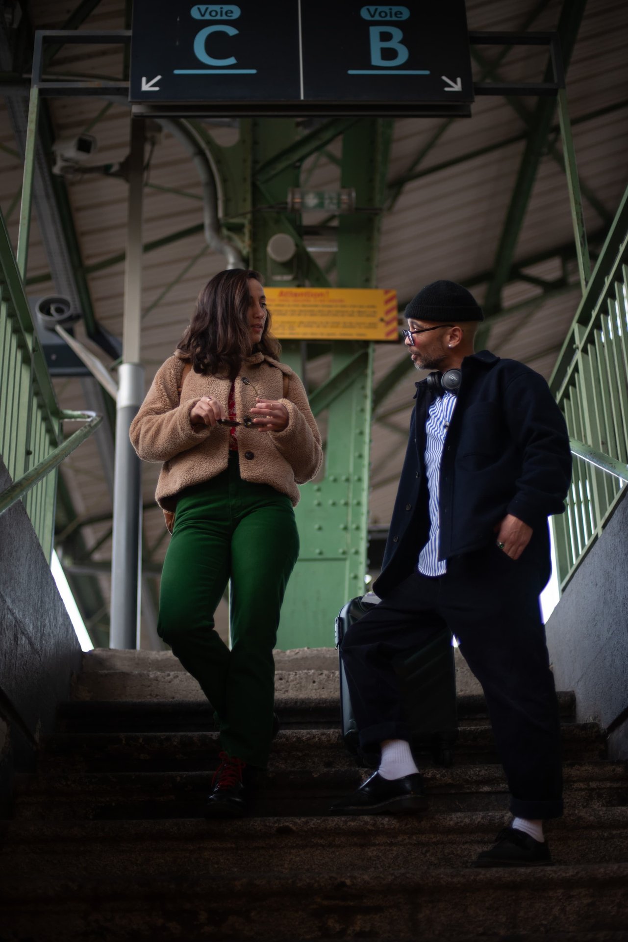 Couple descendant les escaliers d'une gare, panneaux de voies ferroviaires en arrière-plan — séance engagement urbaine Perpignan