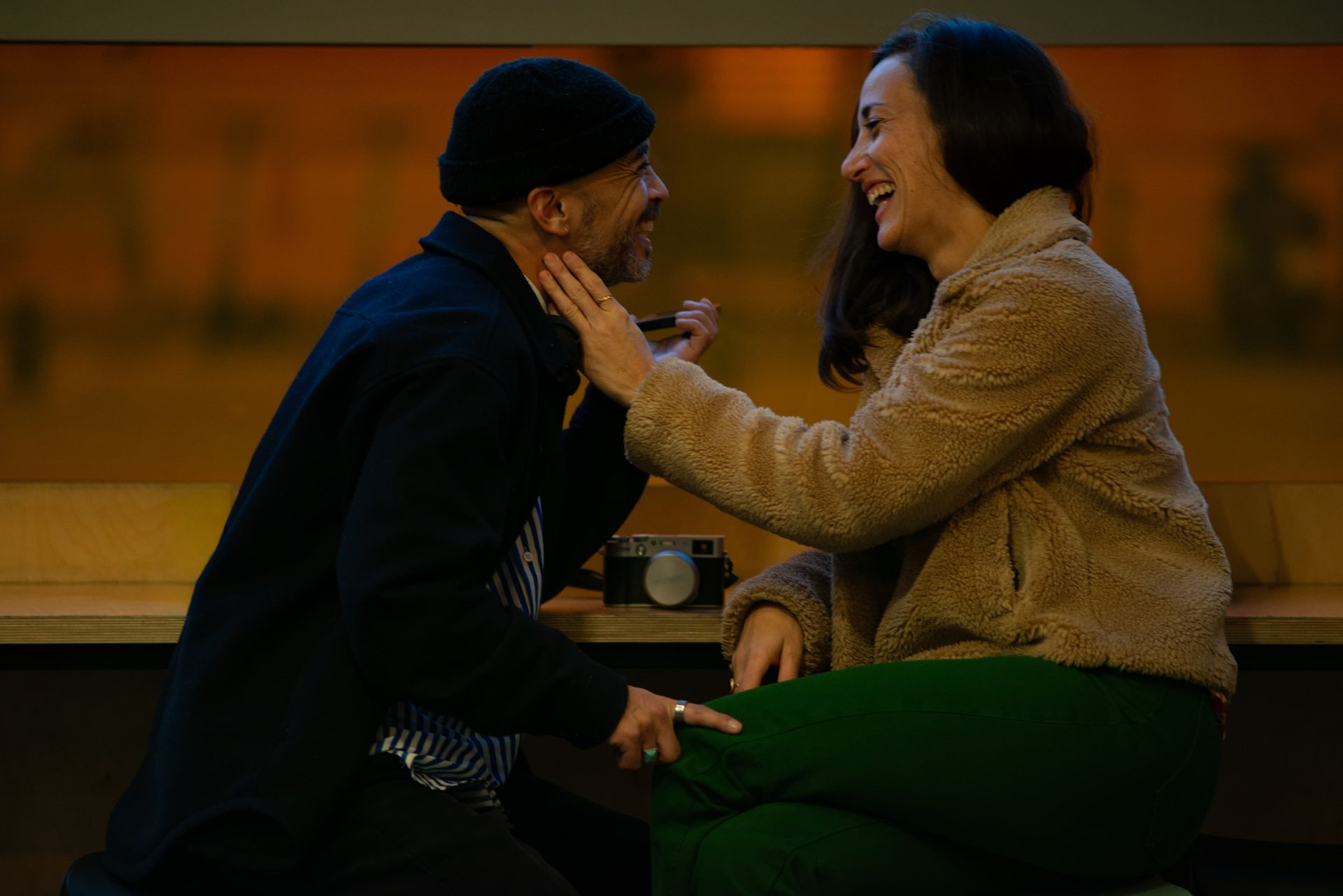 Couple complice face à face dans une lumière ambrée, rires partagés, appareil photo posé entre eux — séance engagement