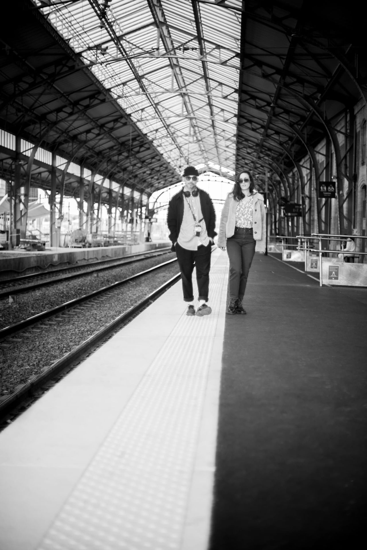 Couple marchant main dans la main sur un quai de gare sous la charpente métallique — séance engagement en noir et blanc