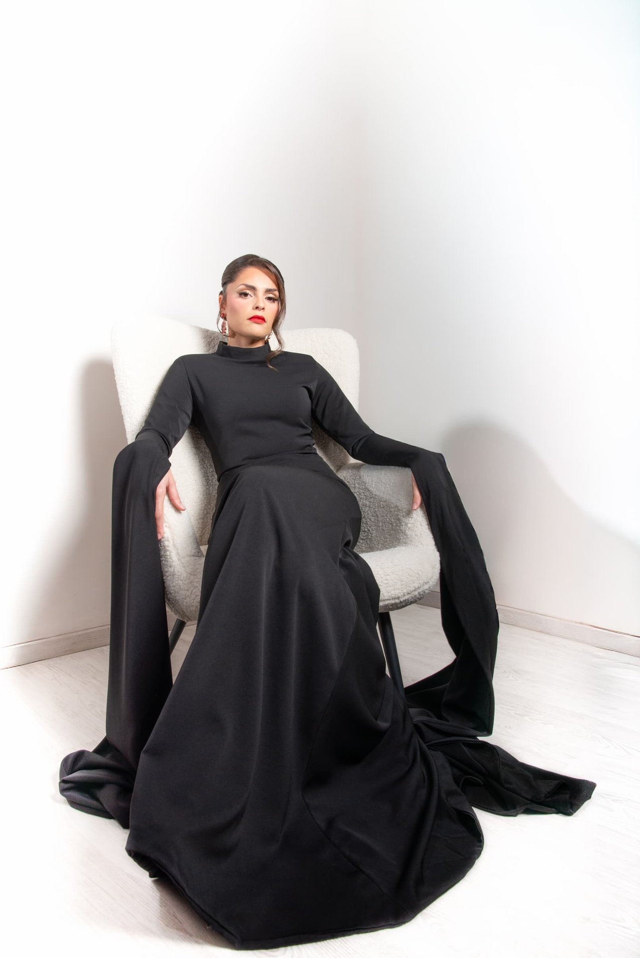 Modèle en robe noire à longue traîne et lèvres rouges, regard intense depuis un fauteuil blanc — shooting inspiration mariage